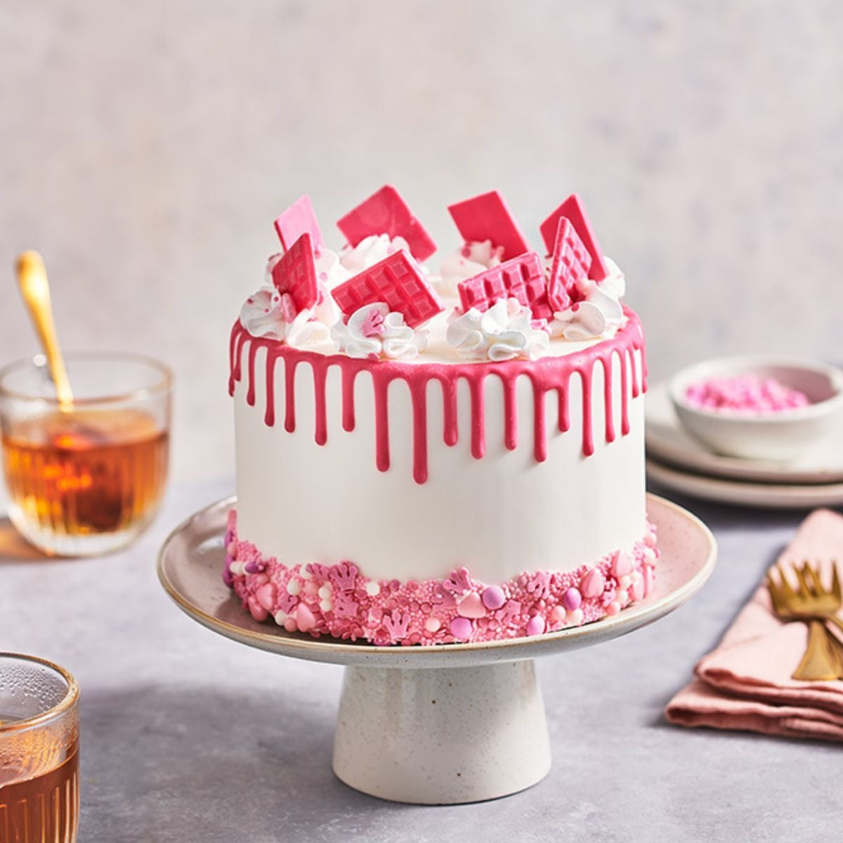 Taart met roze drip, roze chocoladeplakken en roze sprinkles onderaan op een taartplateau