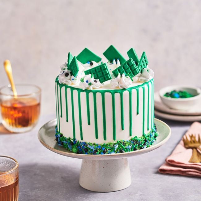 Taart met groene drip, groene chocoladeplakken en blauw-groene sprinkles onderaan op een taartplateau