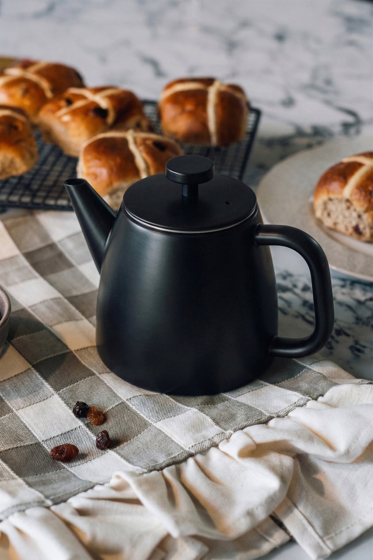 Zwarte theepot met gesloten deksel op een geruite theedoek, met hot cross buns op een rooster en een bord met een broodje erachter
