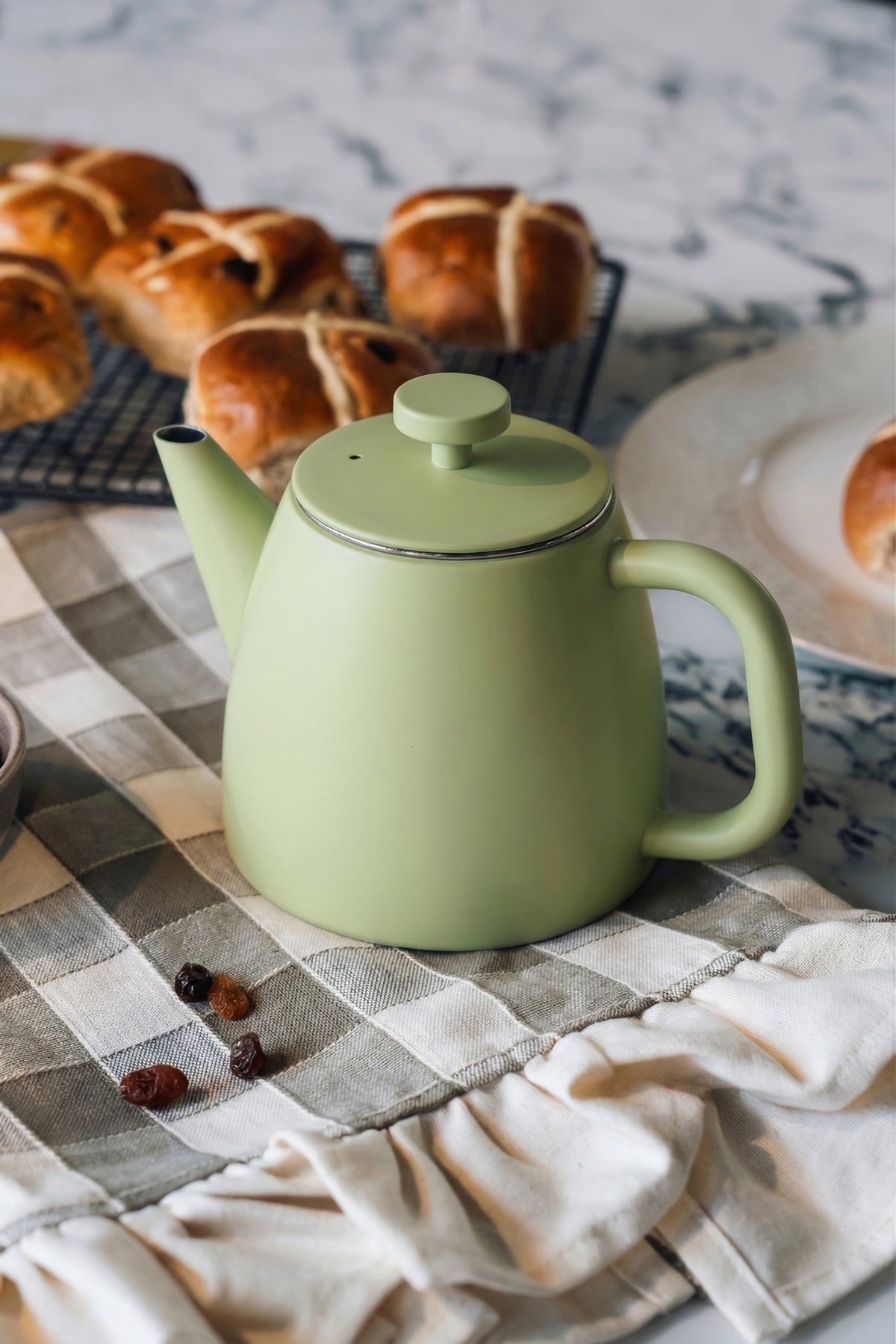 Lichtgroene theepot met gesloten deksel op een grijs geruite theedoek, met hot cross buns op een rooster en een bord erachter