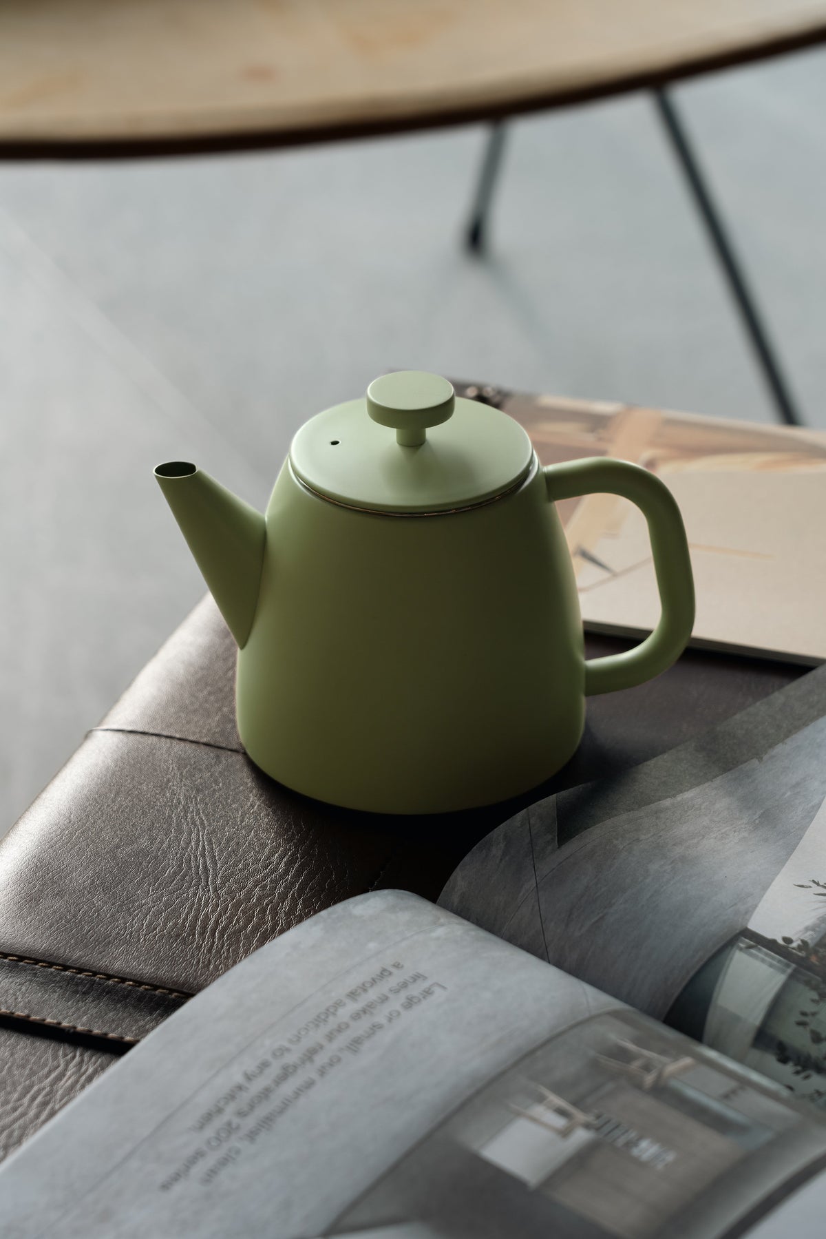 Lichtgroene theepot op een leren bank naast een opengeslagen tijdschrift, met een tafelpoot en houten blad op de achtergrond