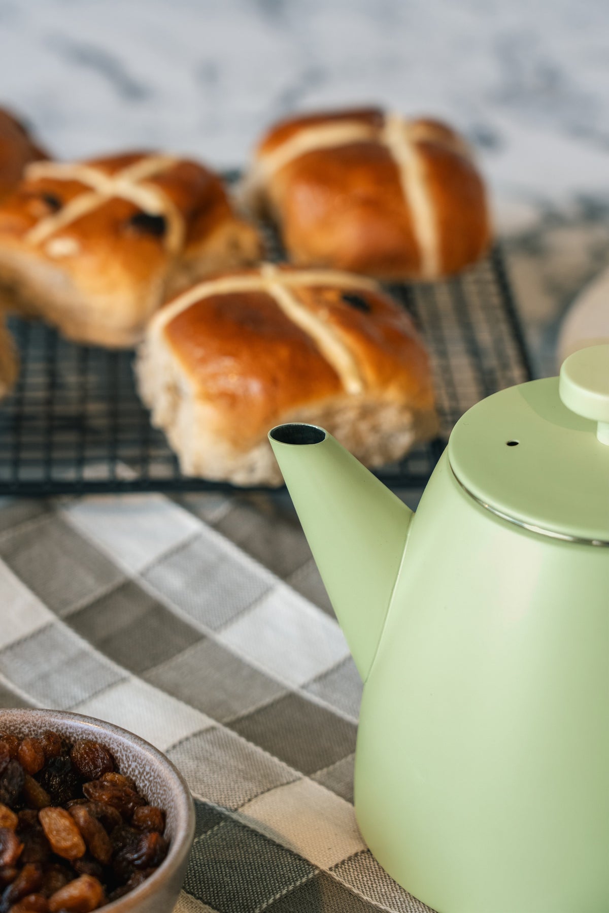 Tuit en deksel van de lichtgroene theepot van dichtbij, met hot cross buns en een kommetje rozijnen onscherp erachter