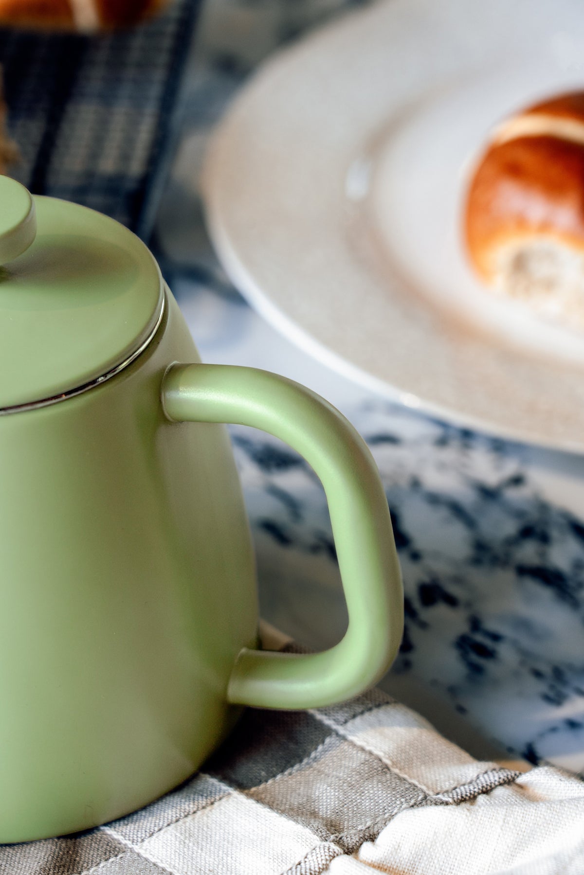 Afgerond handvat en dekselrand van de lichtgroene theepot van dichtbij, met een bord en een broodje op de achtergrond