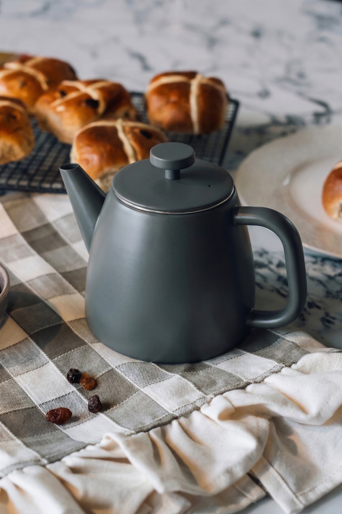 Donkergrijze theepot met gesloten deksel op een grijs geruite theedoek, met hot cross buns op een rooster en een bord erachter