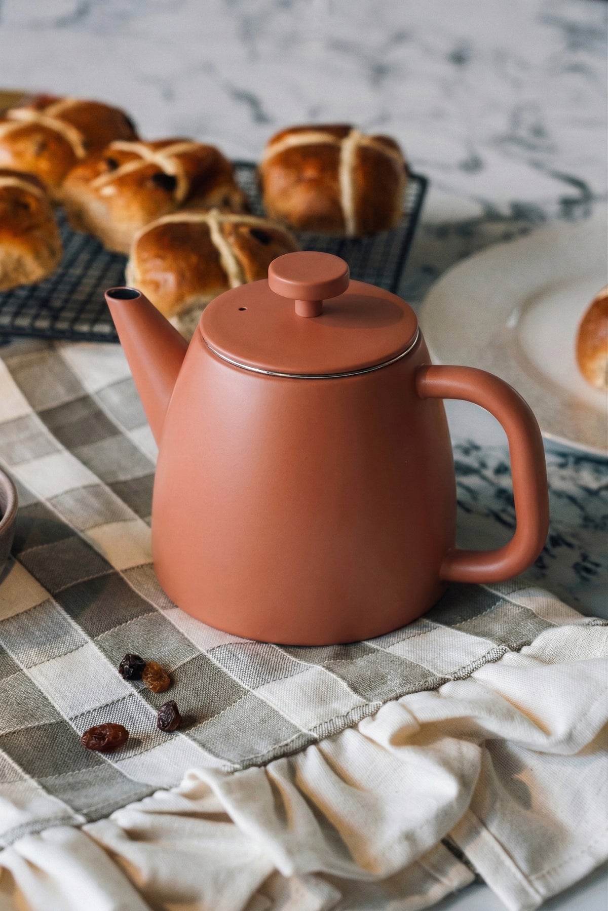 Roodoranje theepot met ronde dekselknop op een geruite theedoek, met hot cross buns en een bord op de achtergrond