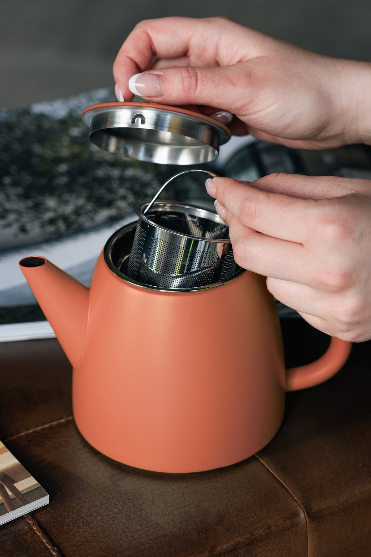 Een hand tilt het deksel op en houdt het metalen zeefmandje boven de opening van de roodoranje theepot