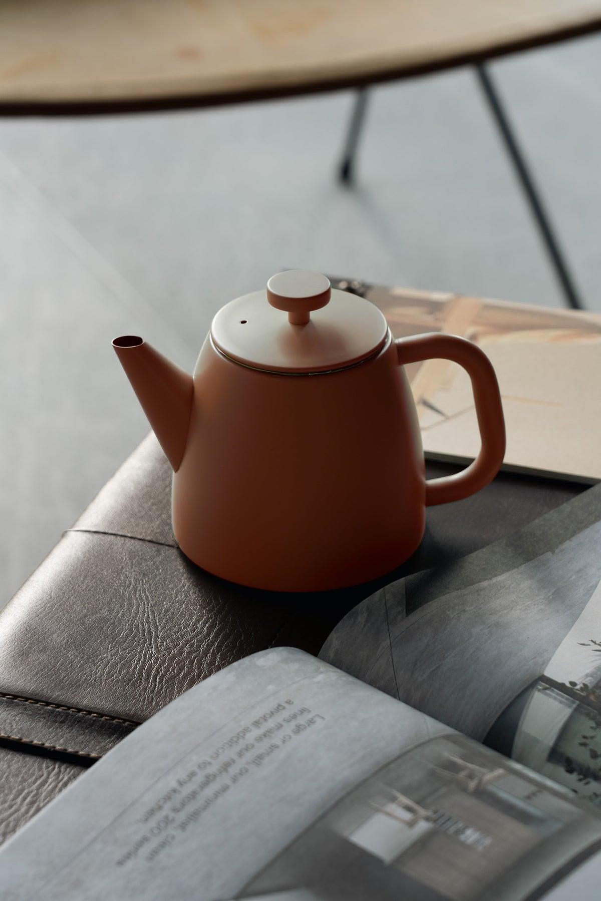 Roodoranje theepot op een leren bank naast een opengeslagen tijdschrift, met een ronde houten tafel erboven