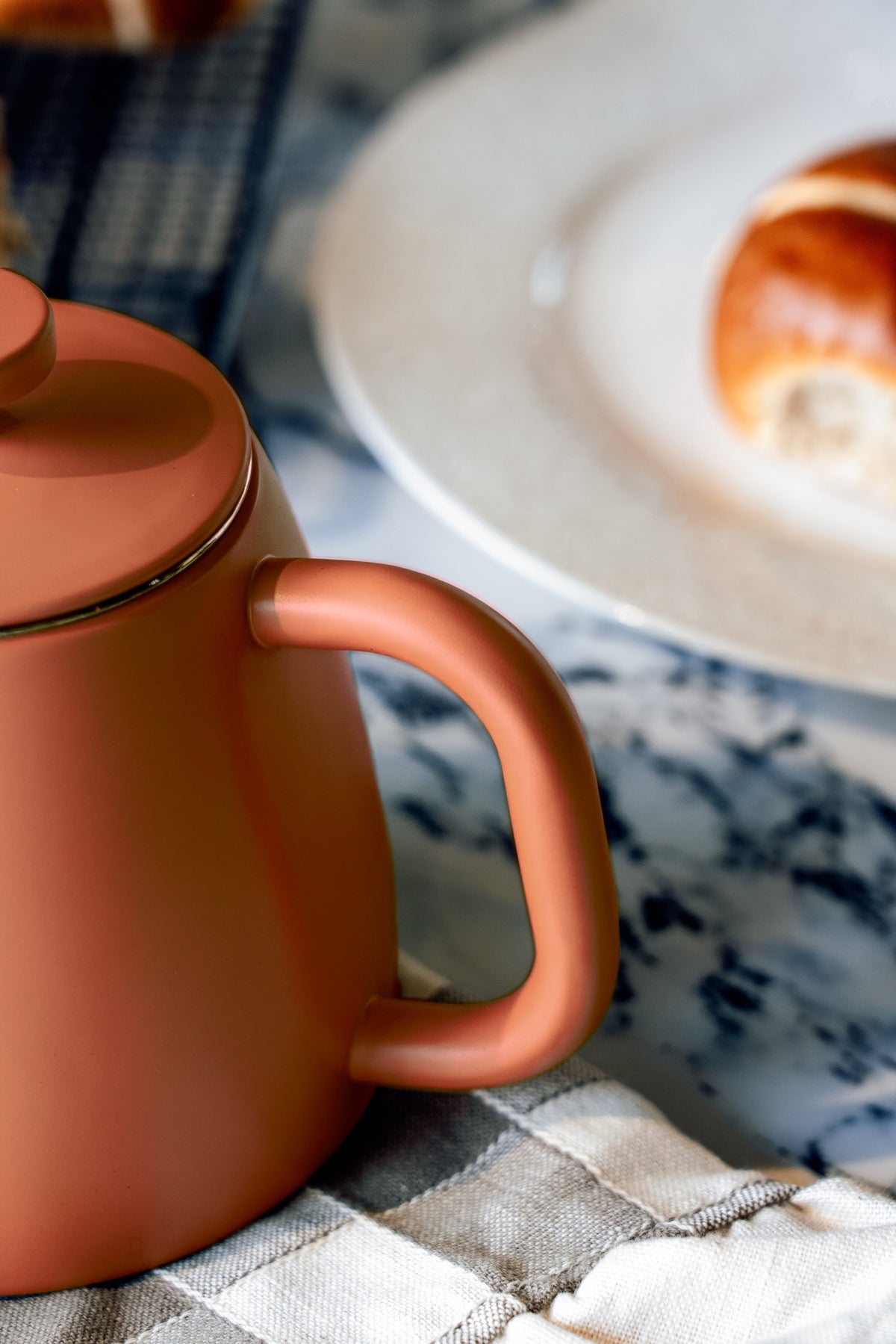 Afgerond handvat van de roodoranje theepot van dichtbij, op een geruite doek naast een bord met een broodje