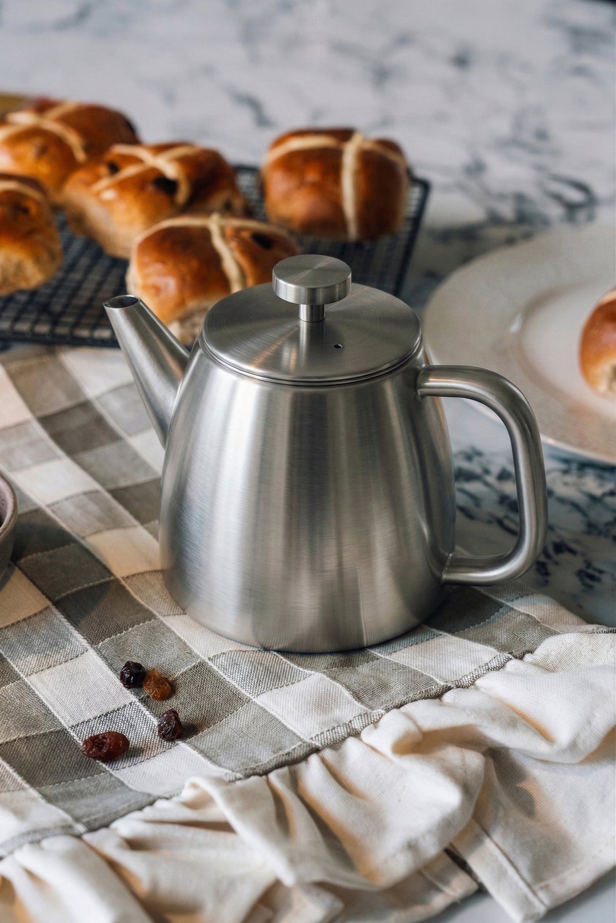 Roestvrijstalen theepot met gesloten deksel op een grijs geruite theedoek, met hot cross buns op een koelrooster erachter