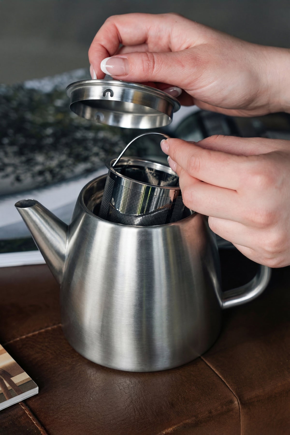 Twee handen tillen het deksel van de roestvrijstalen theepot op en houden het geperforeerde zeefmandje met draadbeugel boven de opening
