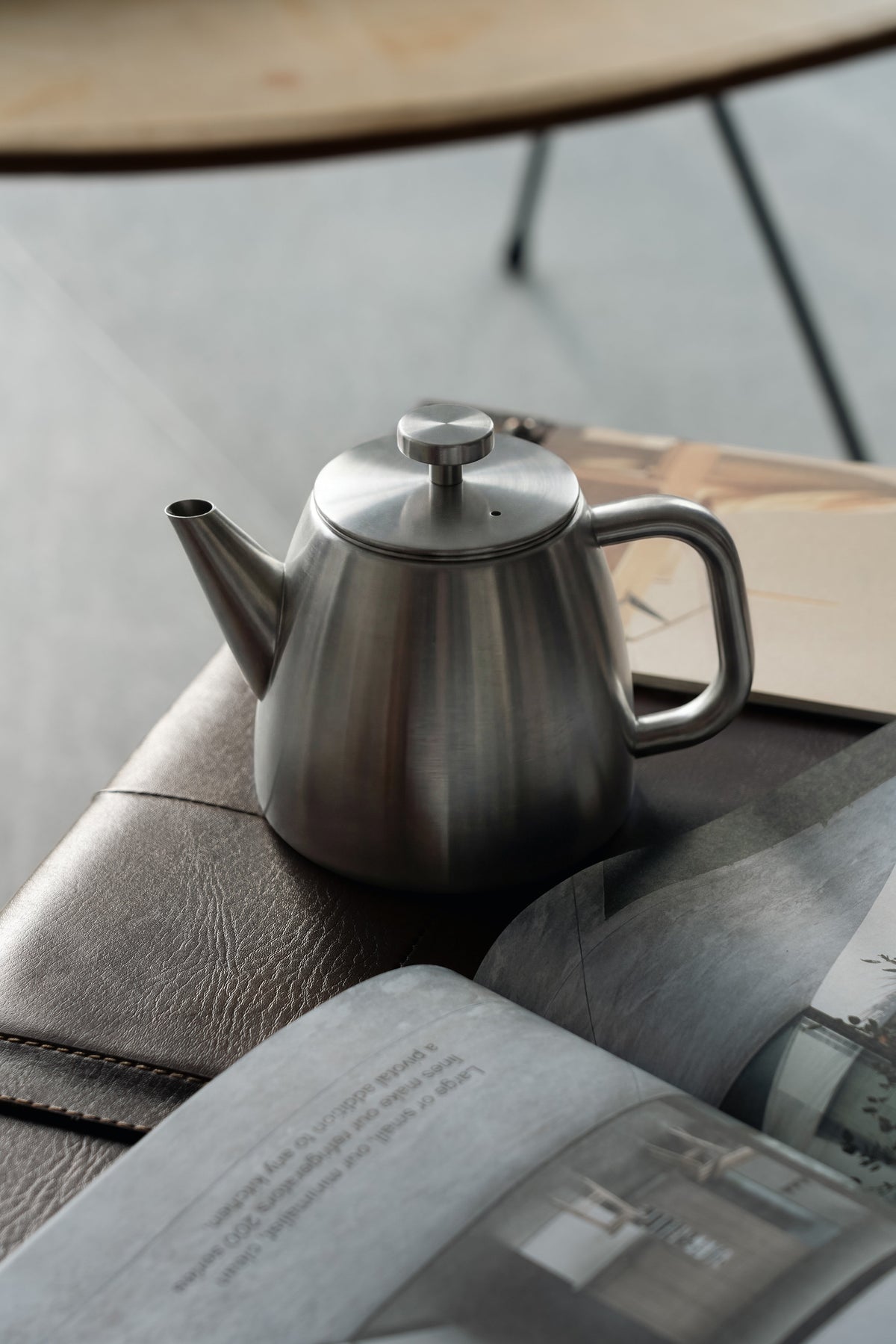 Roestvrijstalen theepot op een leren bank naast een opengeslagen tijdschrift, met een houten tafel op de achtergrond