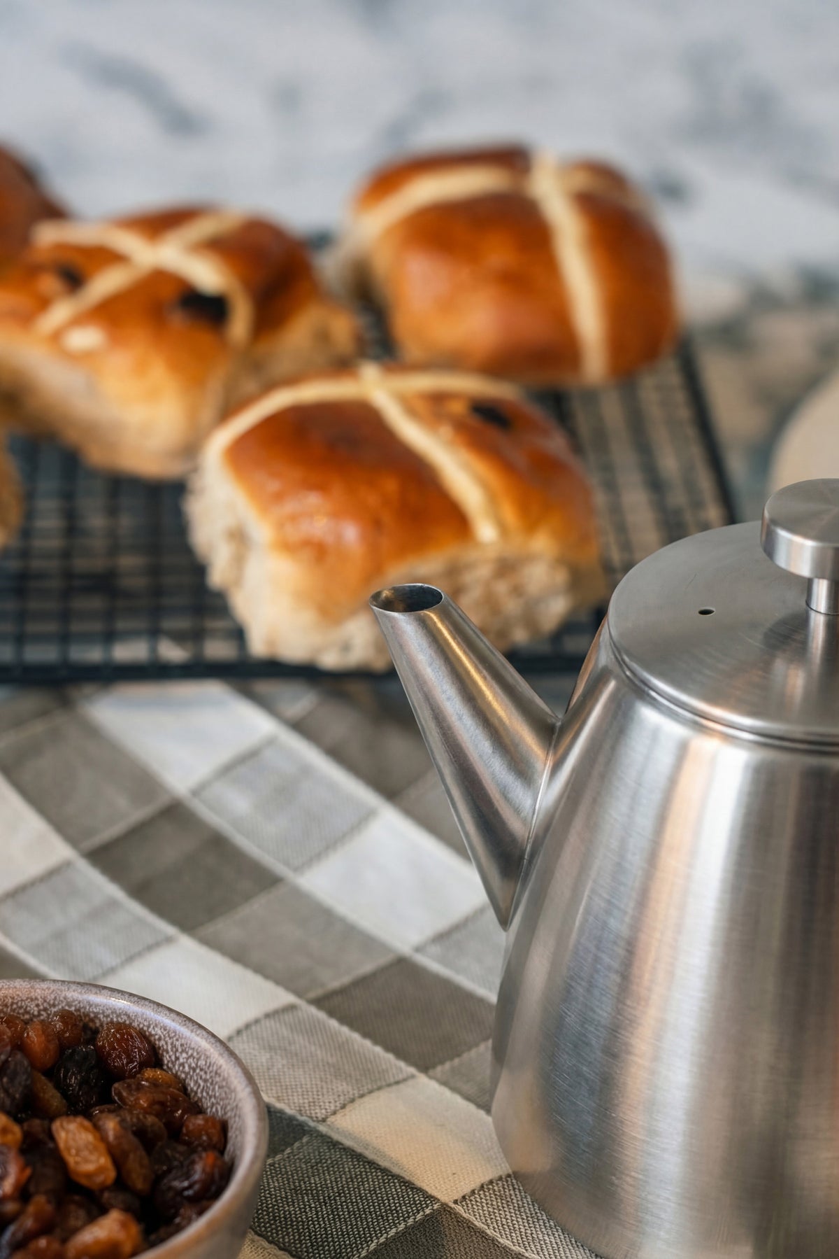 Tuit en dekselrand van de roestvrijstalen theepot van dichtbij, met hot cross buns en een kommetje rozijnen op de achtergrond