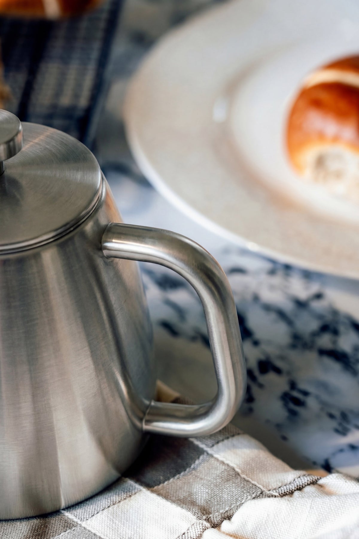 Afgerond handvat en deksel van de roestvrijstalen theepot van dichtbij, met een bord en een broodje wazig erachter