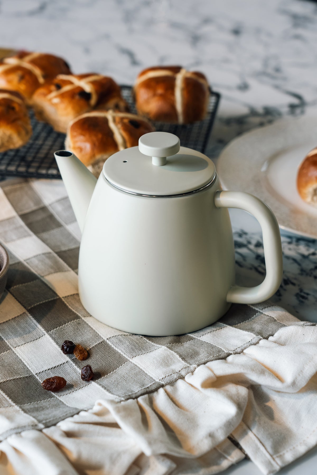 Crèmewitte theepot met ronde dekselknop op een grijs geruite theedoek, met hot cross buns op een rooster en rozijnen op de doek