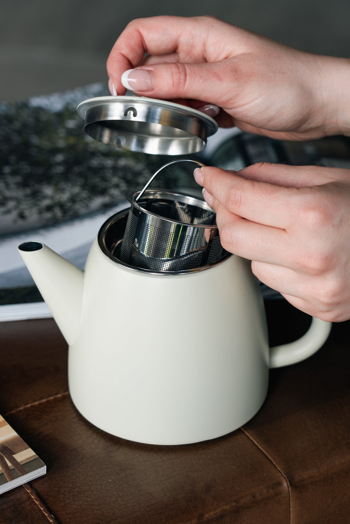 Twee handen tillen het deksel van de crèmewitte theepot op en houden het zeefmandje met draadbeugel boven de opening