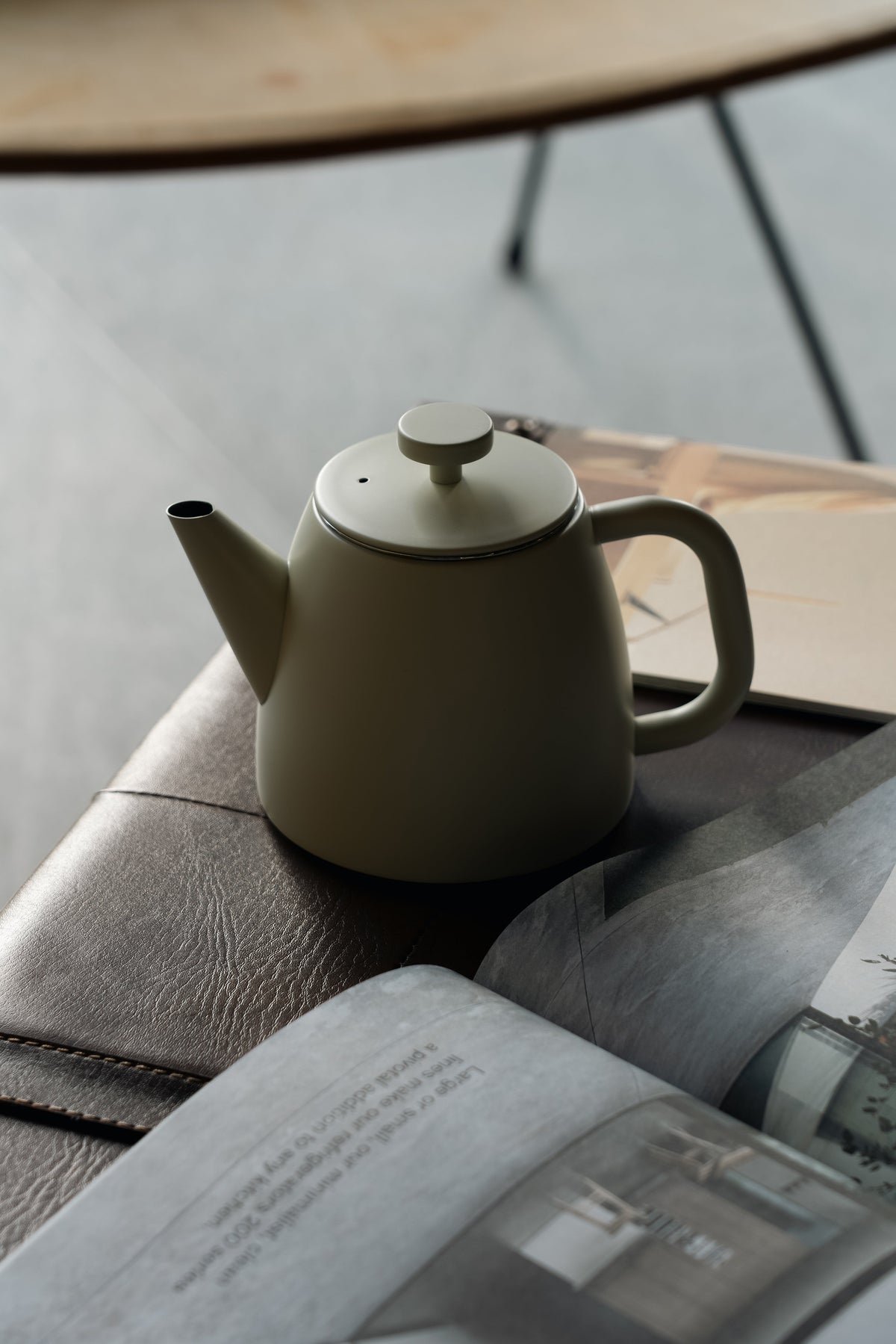 Crèmewitte theepot op een leren bank naast een opengeslagen tijdschrift, met een ronde tafel op de achtergrond