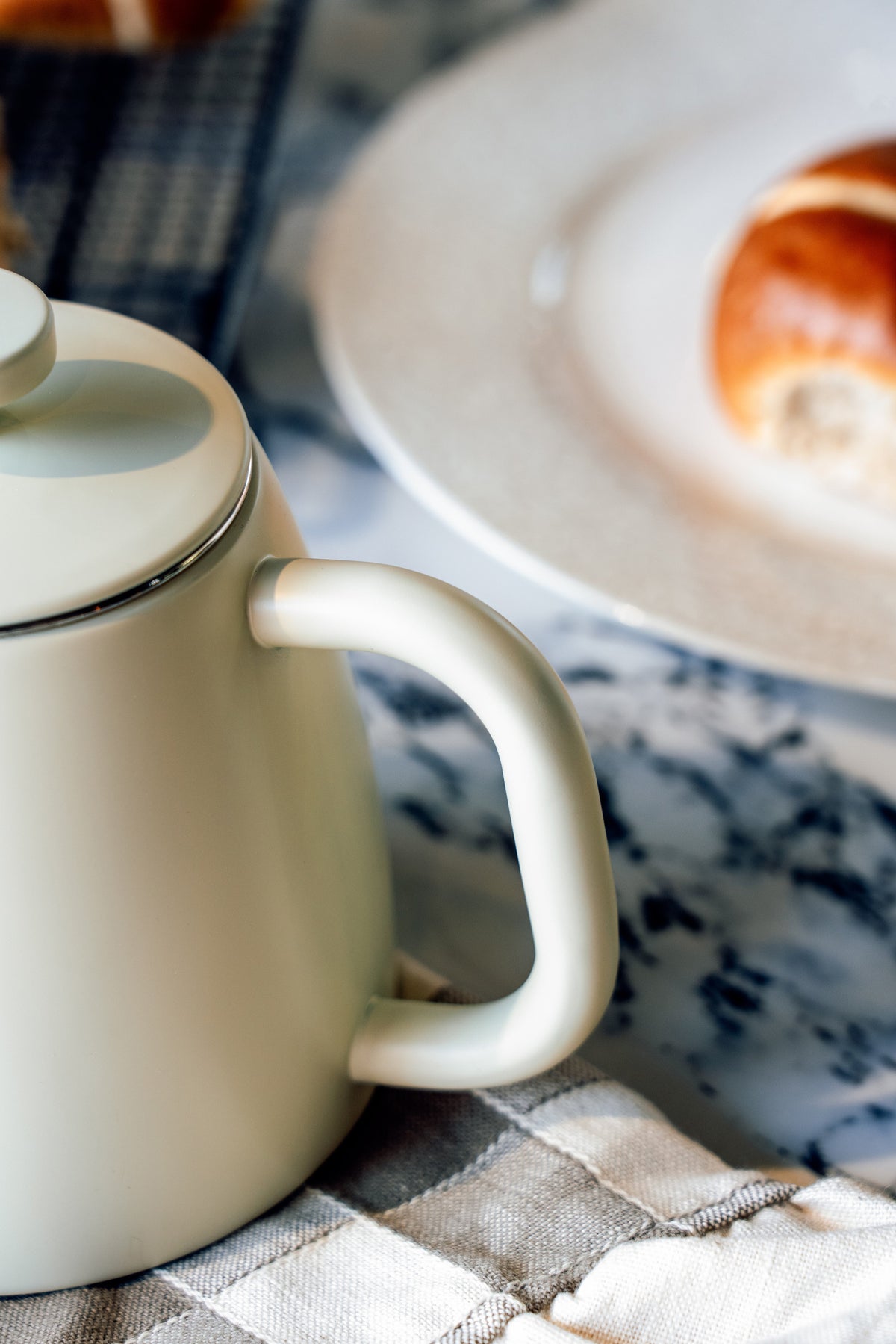 Handvat en dekselrand van de crèmewitte theepot van dichtbij, met een bord met een broodje wazig op de achtergrond