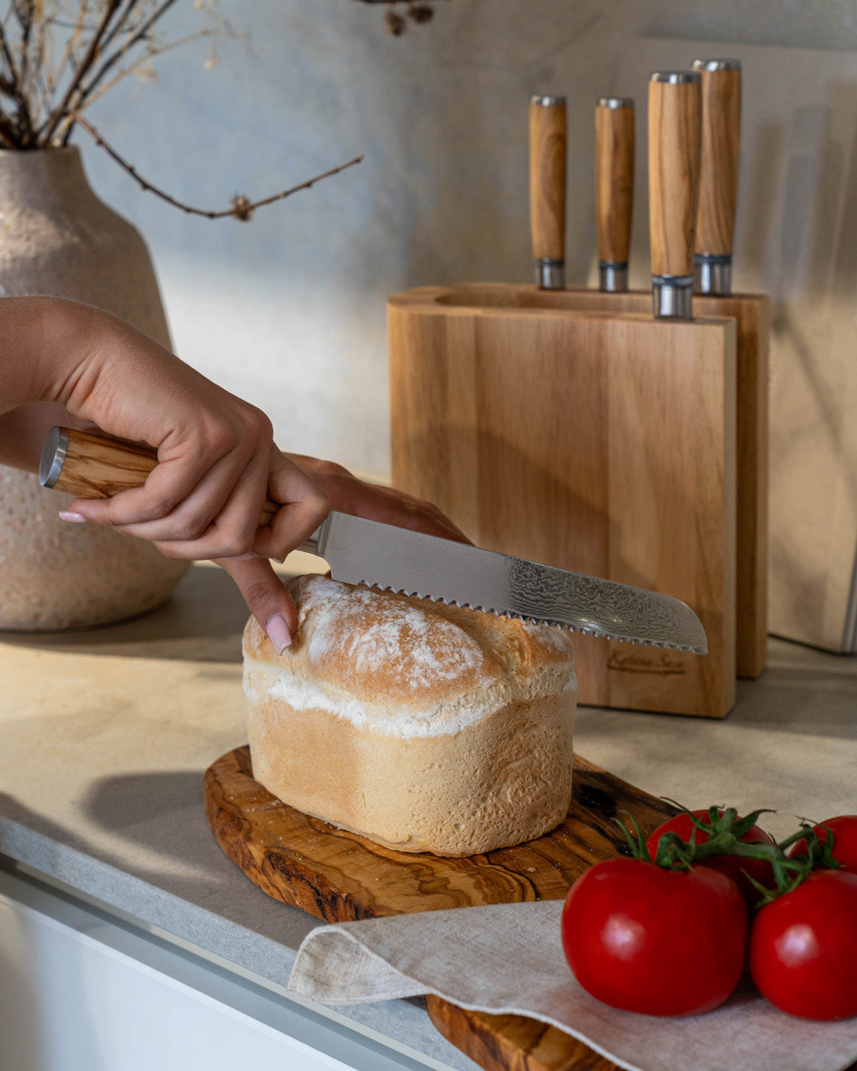 Hand snijdt met een broodmes een brood aan op een olijfhouten plank, met het lichte messenblok op de achtergrond