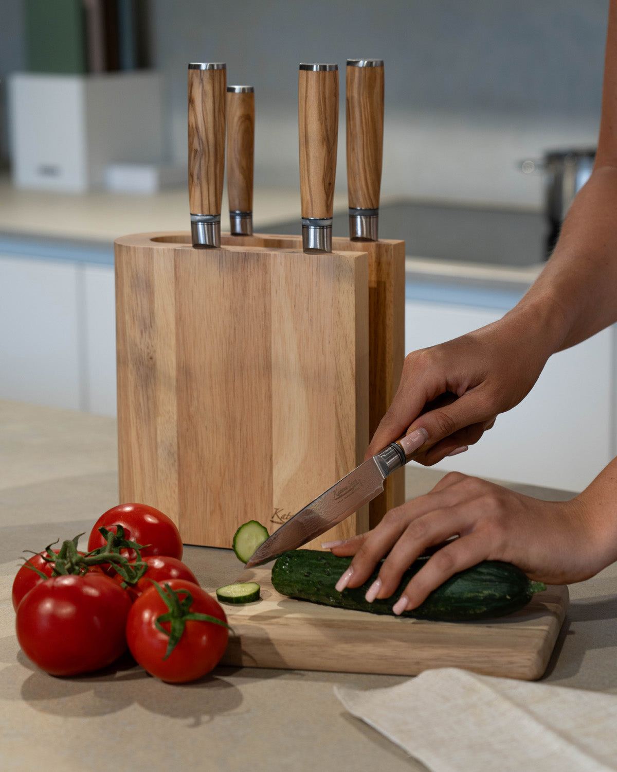 Hand snijdt met een kort mes een komkommer naast tomaten, met het lichte messenblok met vier messen erachter