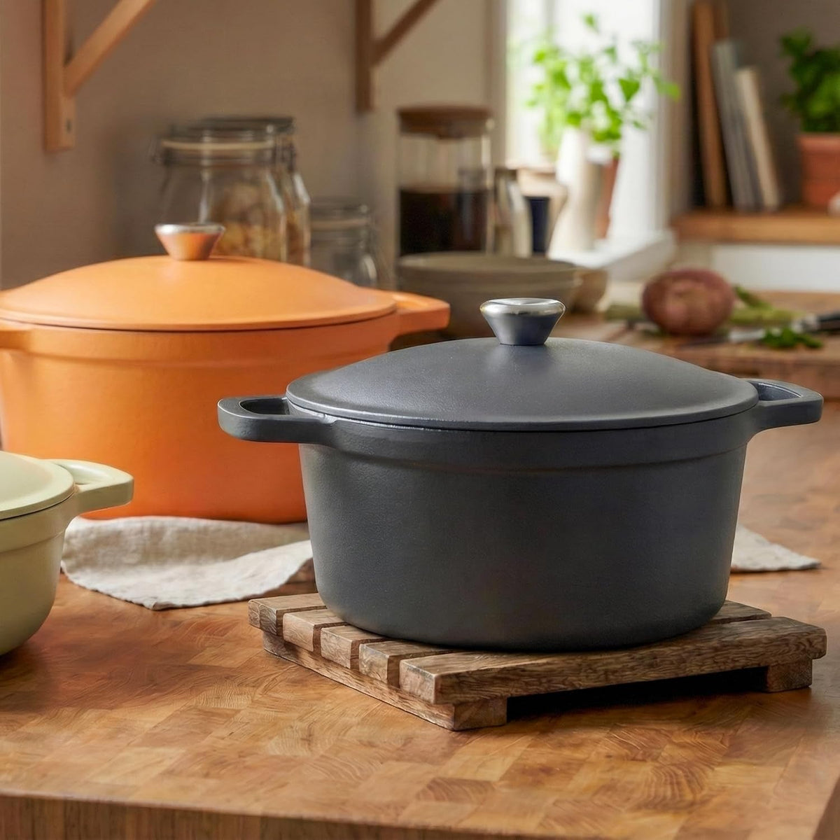 Zwarte braadpan met deksel op een houten onderzetter op een houten aanrecht, met een oranje en een lichtgroene pan op de achtergrond