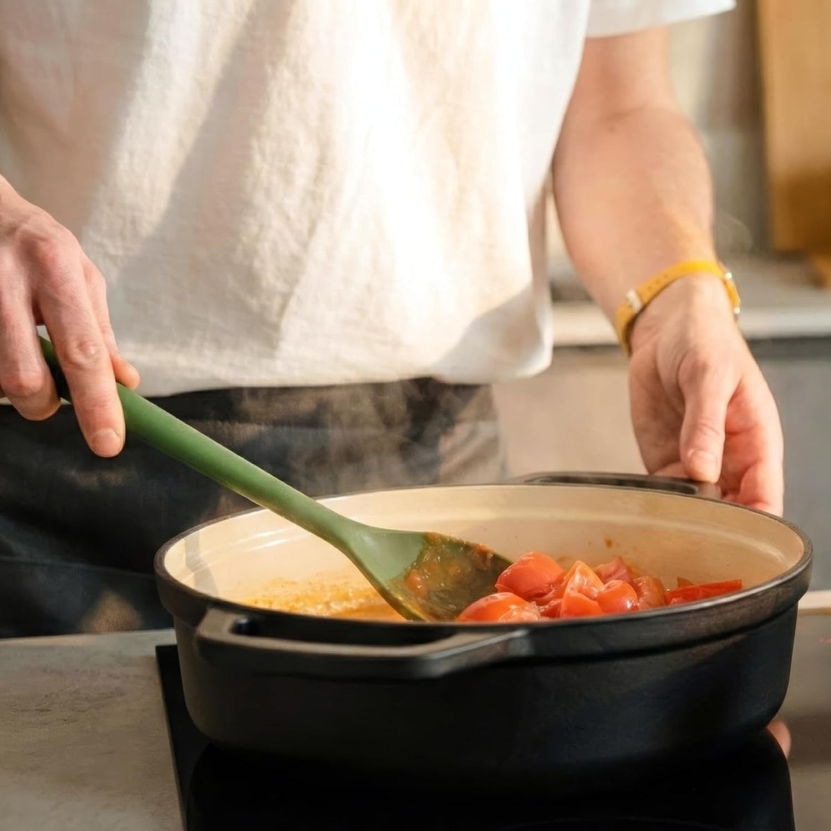 Hand roert met een groene lepel door tomatenstukjes in een lage zwarte braadpan met een crèmekleurige emaille binnenkant op een kookplaat
