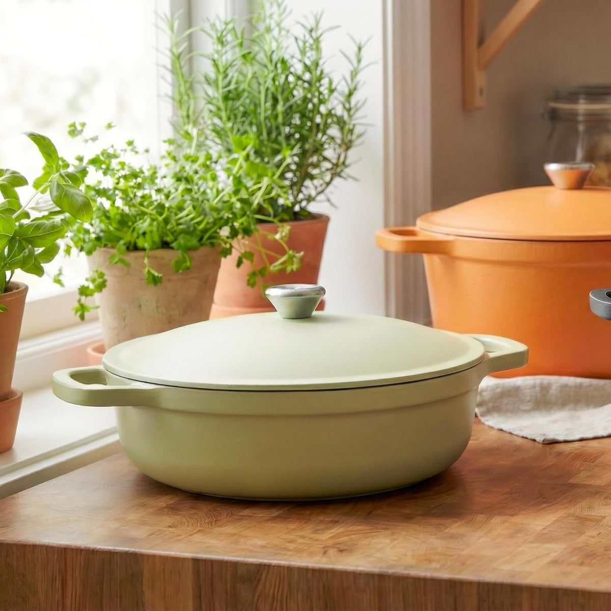 Lichtgroene lage braadpan met deksel op een houten aanrecht voor een raam met kruidenplanten, met een oranje pan op de achtergrond