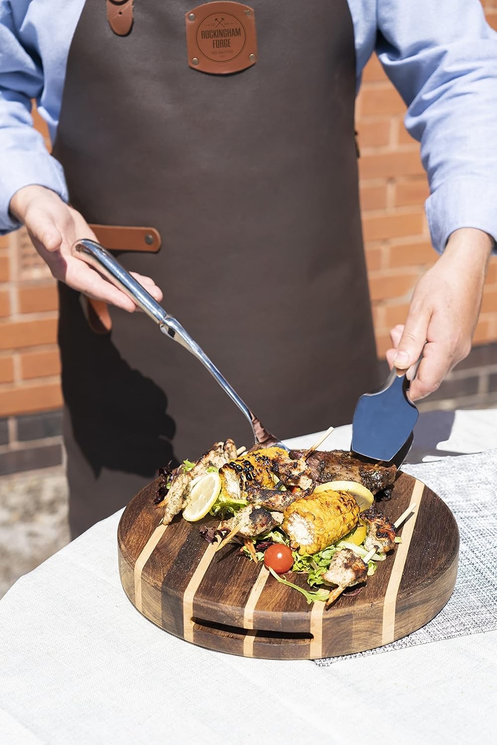 Een man in een bruine schort met opdruk Rockingham Forge serveert met een vork en een spatel gegrilde kip, maïs, citroen en sla van een rond snijblok op een buitentafel