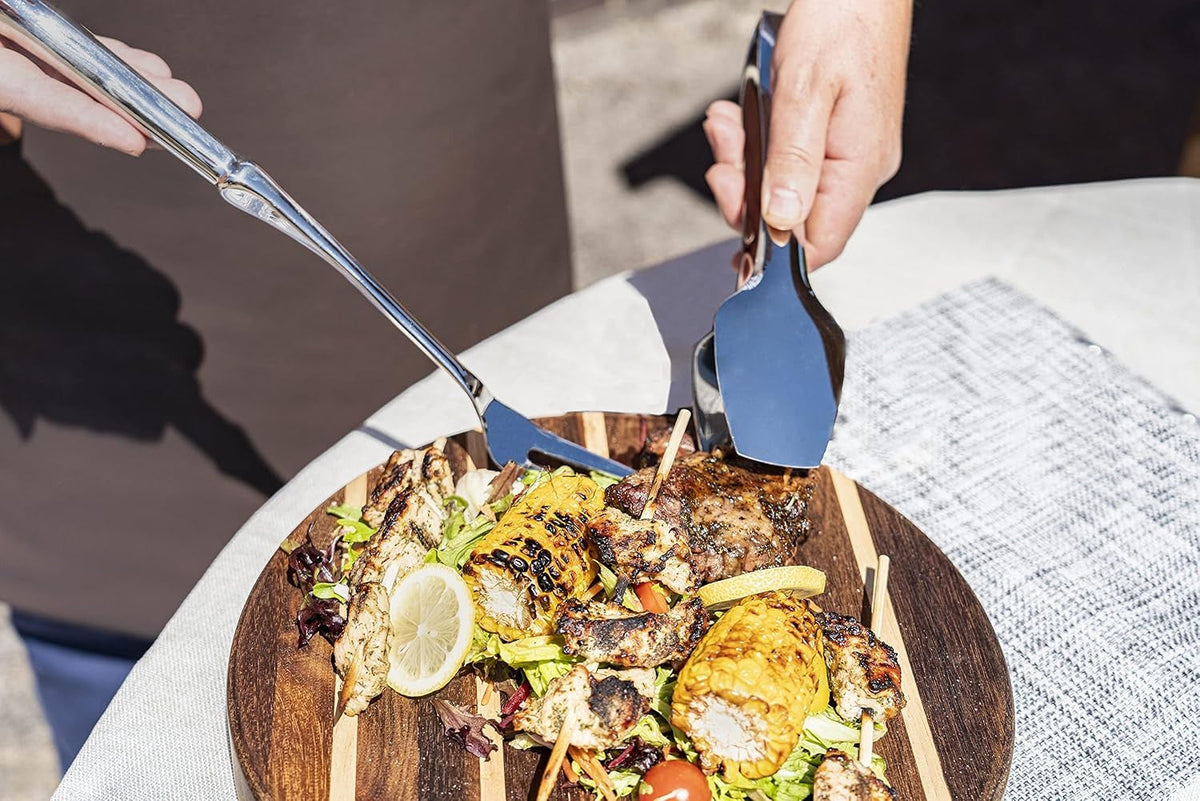 Twee handen met serveerbestek bij een rond houten blok vol gegrilde kip, maïs, citroen en steak op een tafel buiten
