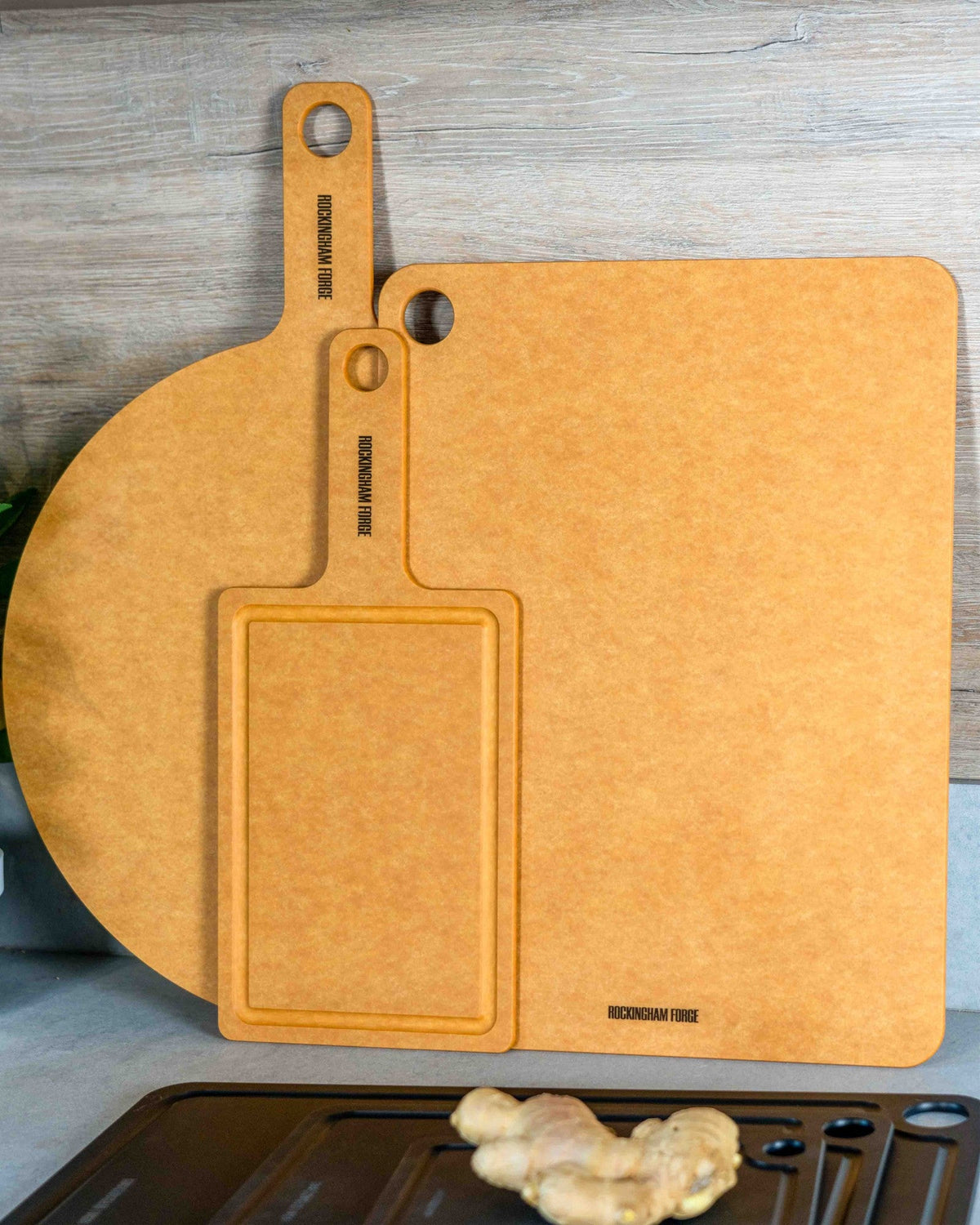 Naturel vezelhouten borden tegen een houten wand: links een groot rond bord met handvat, voor het midden een rechthoekig bord met handvat en rand, rechts een groot vierkant bord met ophangoog