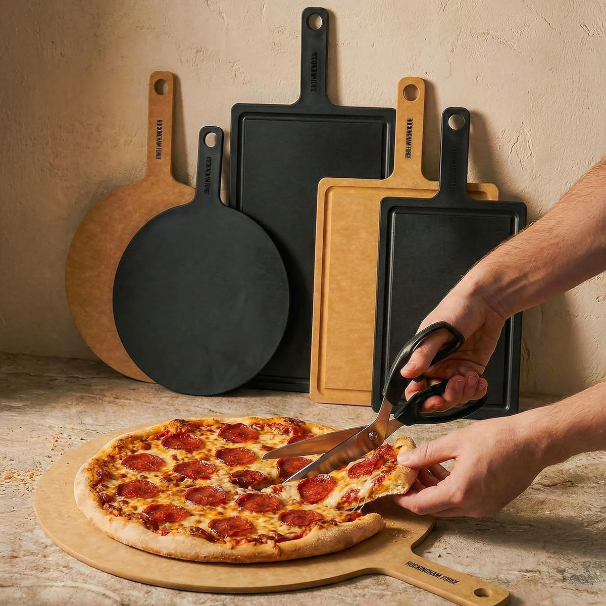 Een hand knipt met een schaar een pepperonipizza in stukken op een naturel bord met handvat, achter het bord staan ronde en rechthoekige borden in naturel en zwart tegen een gepleisterde muur