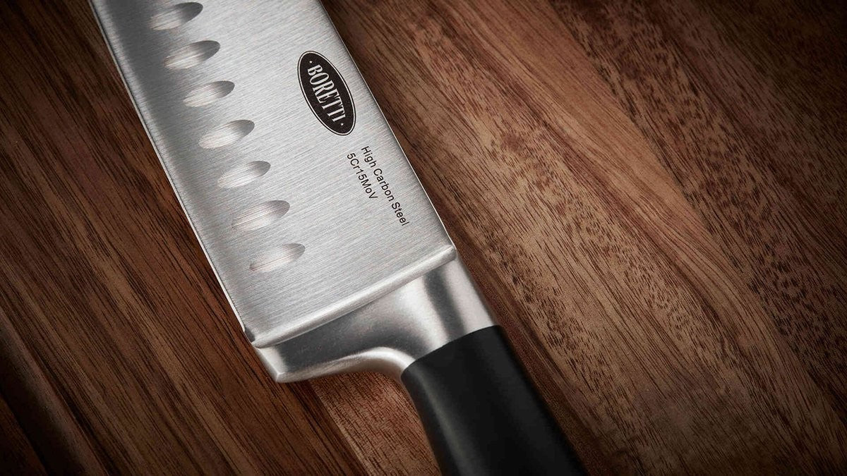 Close-up van het brede lemmet van het Boretti santokumes met kuiltjes en de opdruk 'Boretti High Carbon Steel 5Cr15MoV', op een houten snijplank.