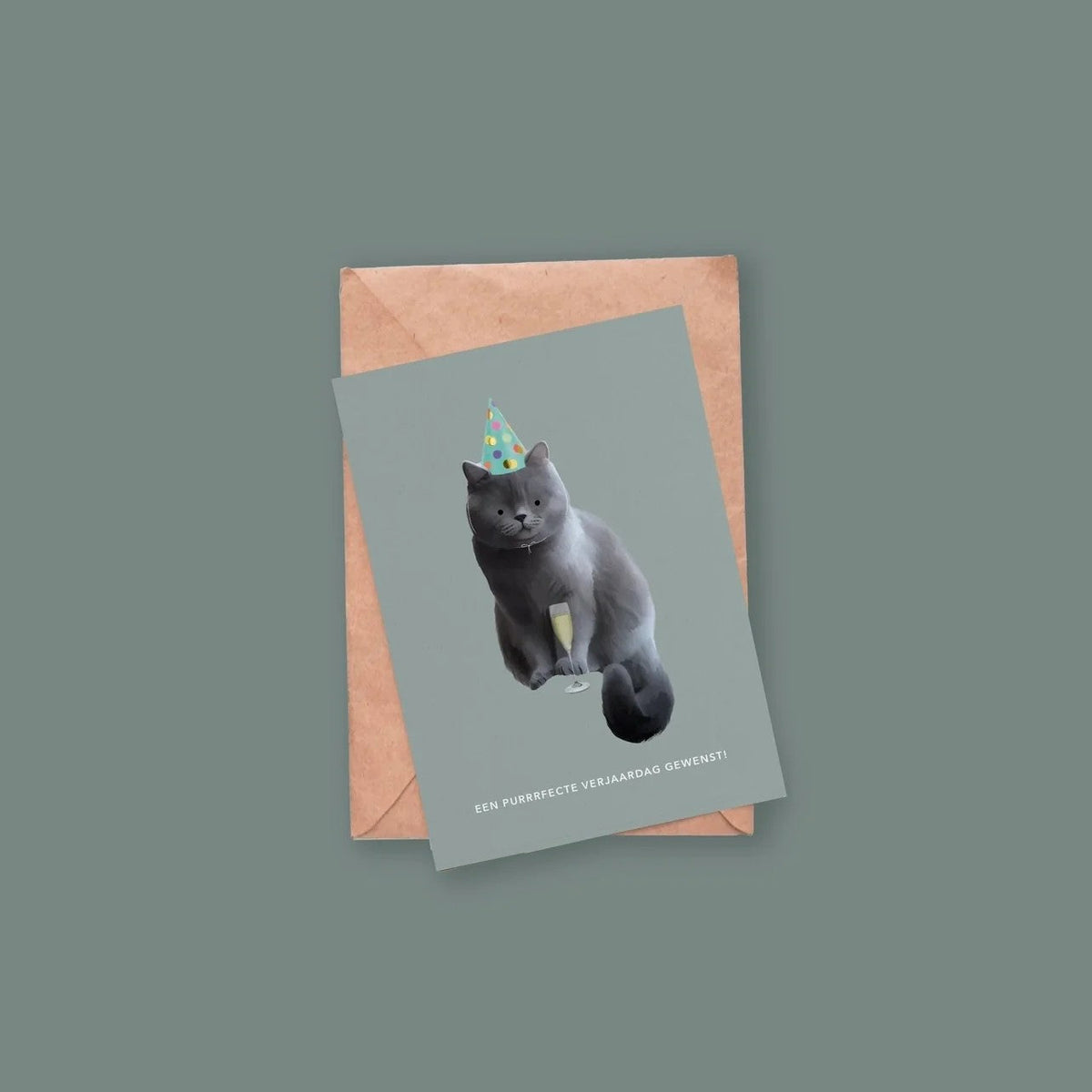 Wenskaart met een kat in feesthoedje en glas bubbels, plat neergelegd op een kraftpapieren envelop tegen een groengrijze achtergrond