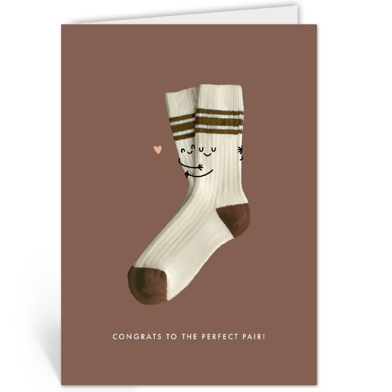 Terracotta wenskaart met knuffelende sokken-illustratie, liggend op een kraftpapieren envelop tegen een bruine achtergrond.