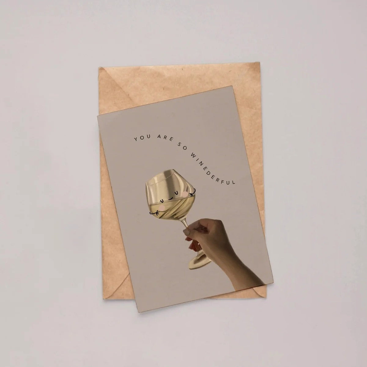 Wenskaart met een lachend wijnglas vastgehouden door een hand, plat neergelegd op een kraftpapieren envelop tegen een grijze achtergrond