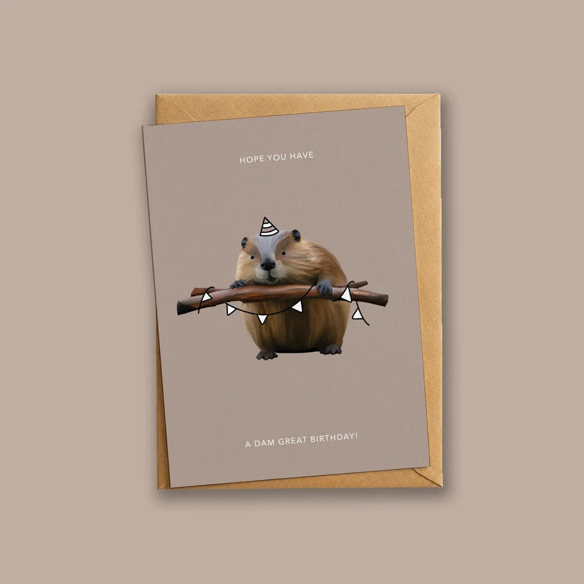 Wenskaart 'Have A Dam Great Birthday' van Depre Design op taupe achtergrond, met een illustratie van een bever met puntmuts die een tak met vlaggenlijn vasthoudt, kraftpapieren envelop eronder.