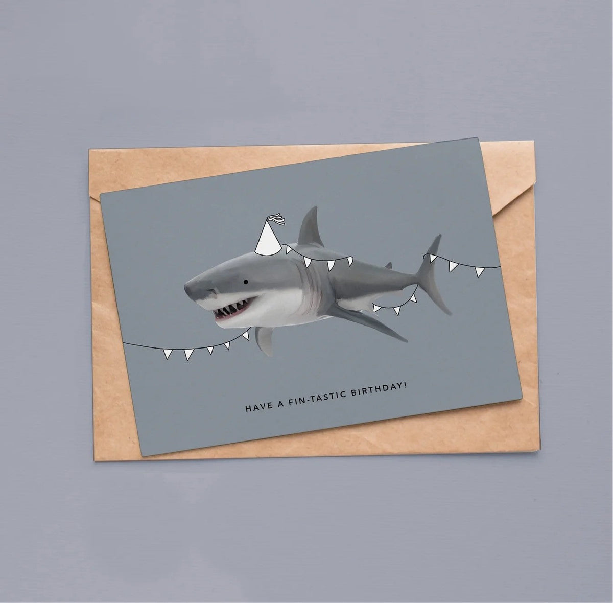 Wenskaart van Depre Design met een geïllustreerde haai met feestmutsje en vlaggenlijn, op een kraft envelop tegen een grijsblauwe achtergrond.