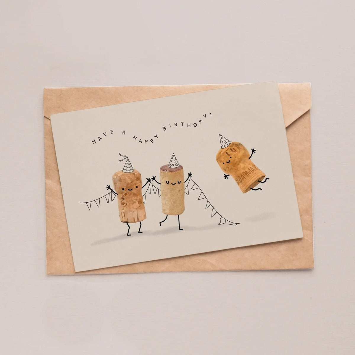 Wenskaart 'Have a happy birthday' van Depre Design met drie dansende kurken in feestmutsjes en een vlaggenlijn, op een kraft envelop.