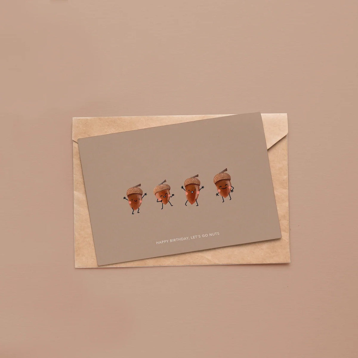 Wenskaart met eikeltjes-illustratie liggend op een kraft envelop tegen roze-beige achtergrond