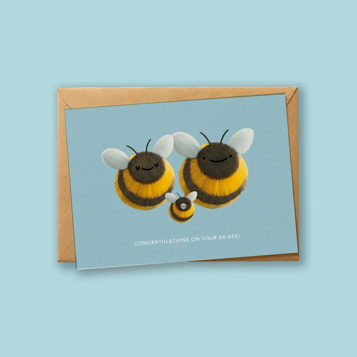 Wenskaart 'Congratulations On Your Ba-Bee' van Depre Design op lichtblauwe achtergrond, met een illustratie van twee grote hommels en een klein babybijtje ertussen, kraftpapieren envelop eronder.