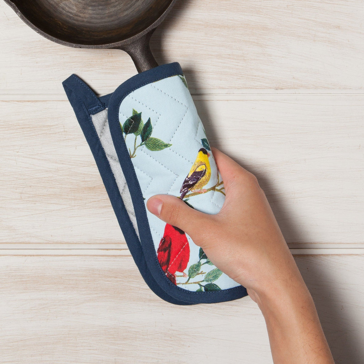 Hand pakt met een pannenlap met vogels het handvat van een gietijzeren koekenpan vast