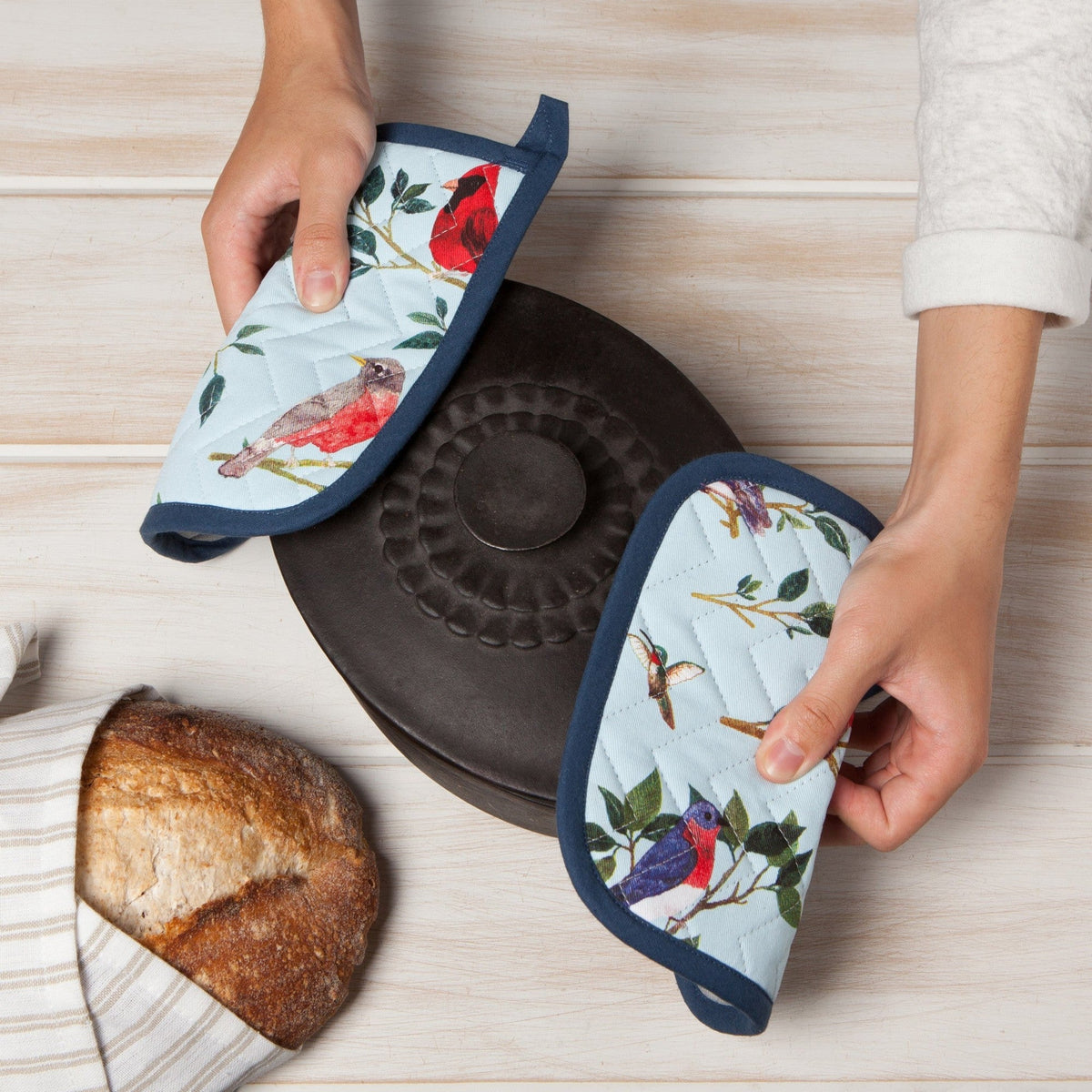 Twee handen houden twee gevouwen pannenlappen vast rond een gietijzeren braadpan, naast een brood in een theedoek