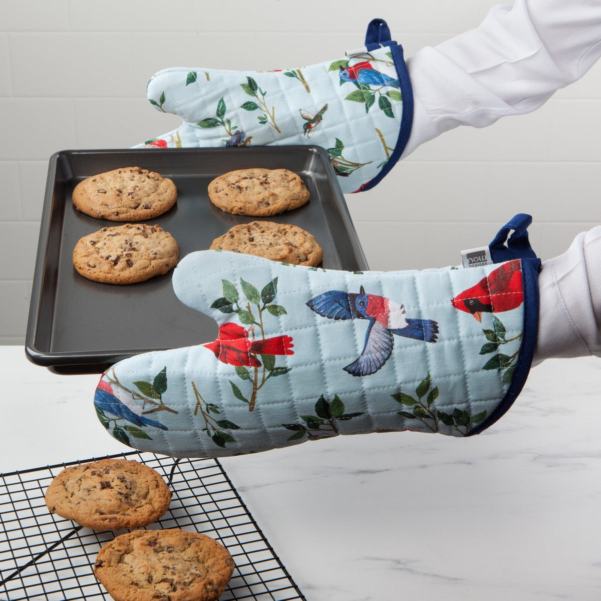 Twee handen in witte mouwen houden met ovenwanten een bakplaat met koekjes boven een afkoelrek