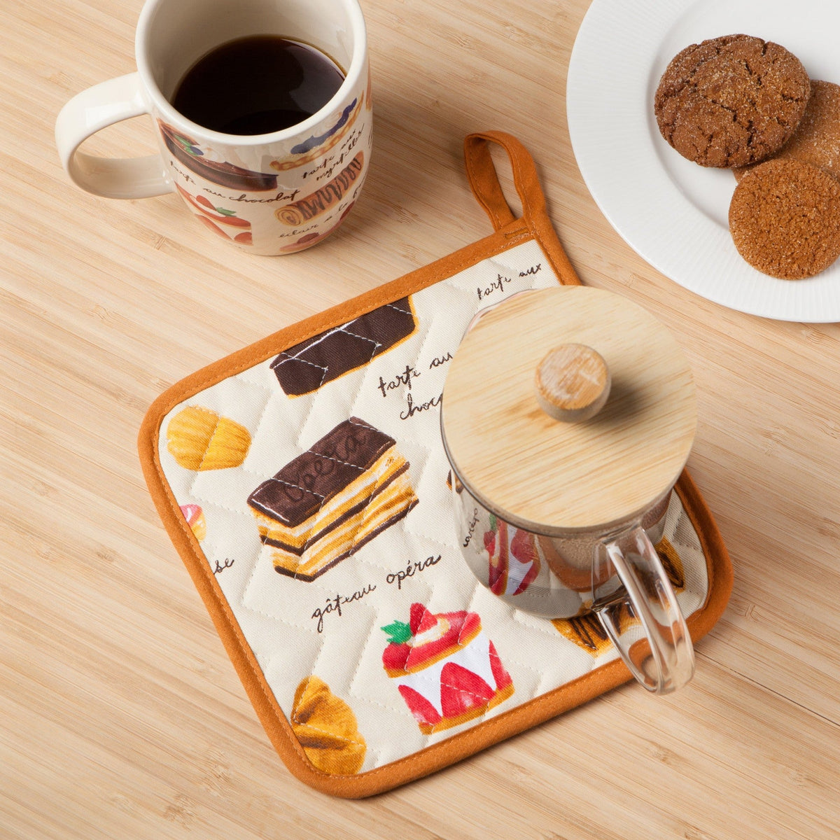 Pannenlap met gebakjes onder een glazen cafetière met bamboedeksel, naast een kop koffie en koekjes op een houten plank