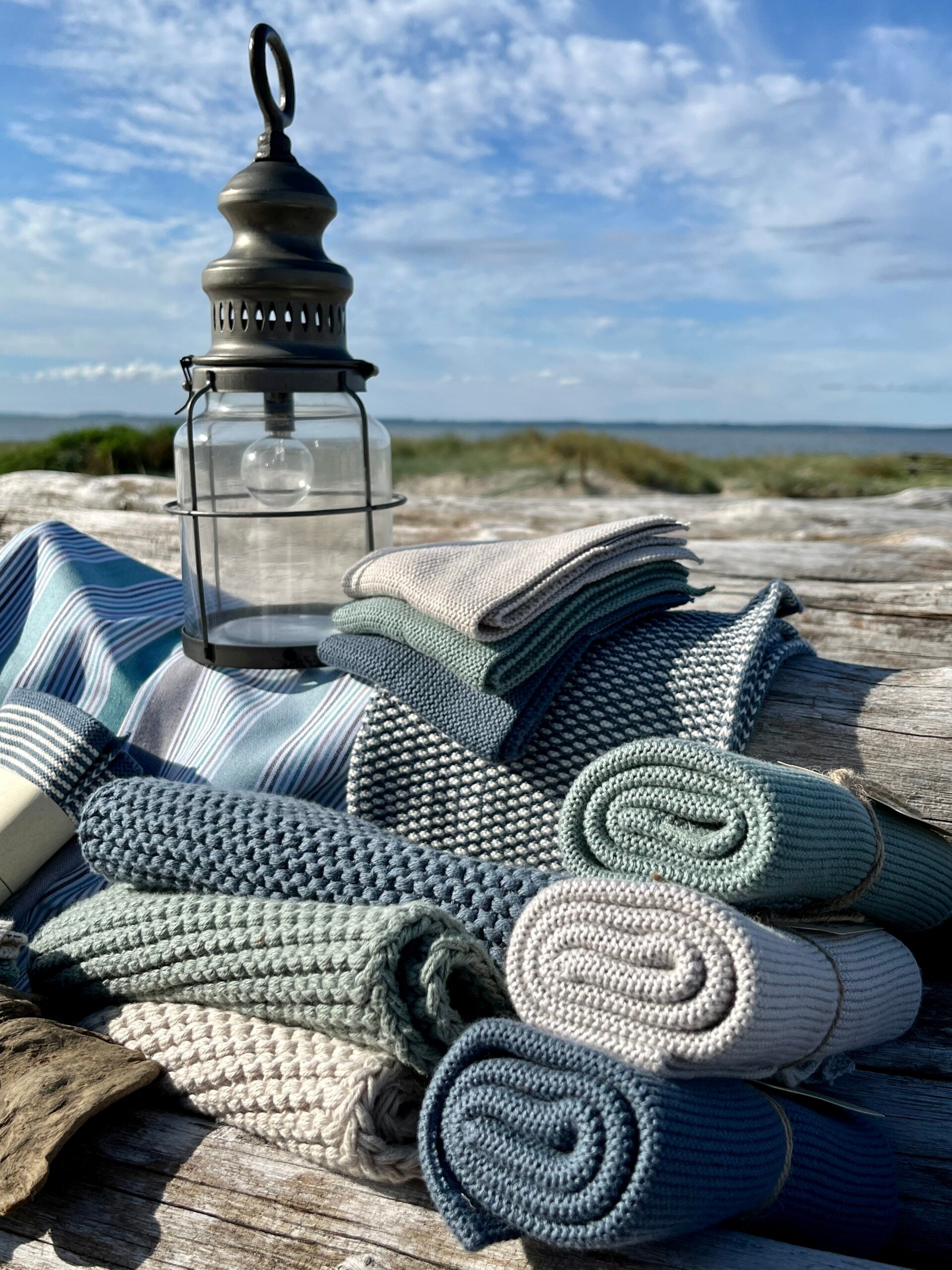 Solwang Design Pannenlap set van 2 - Rustiek Blauw sfeerfoto op verweerd hout aan het strand met een metalen lantaarn, blauw-wit gestreepte stof op de achtergrond en meerdere opgerolde geribde doeken in blauwtinten, grijs en crème.