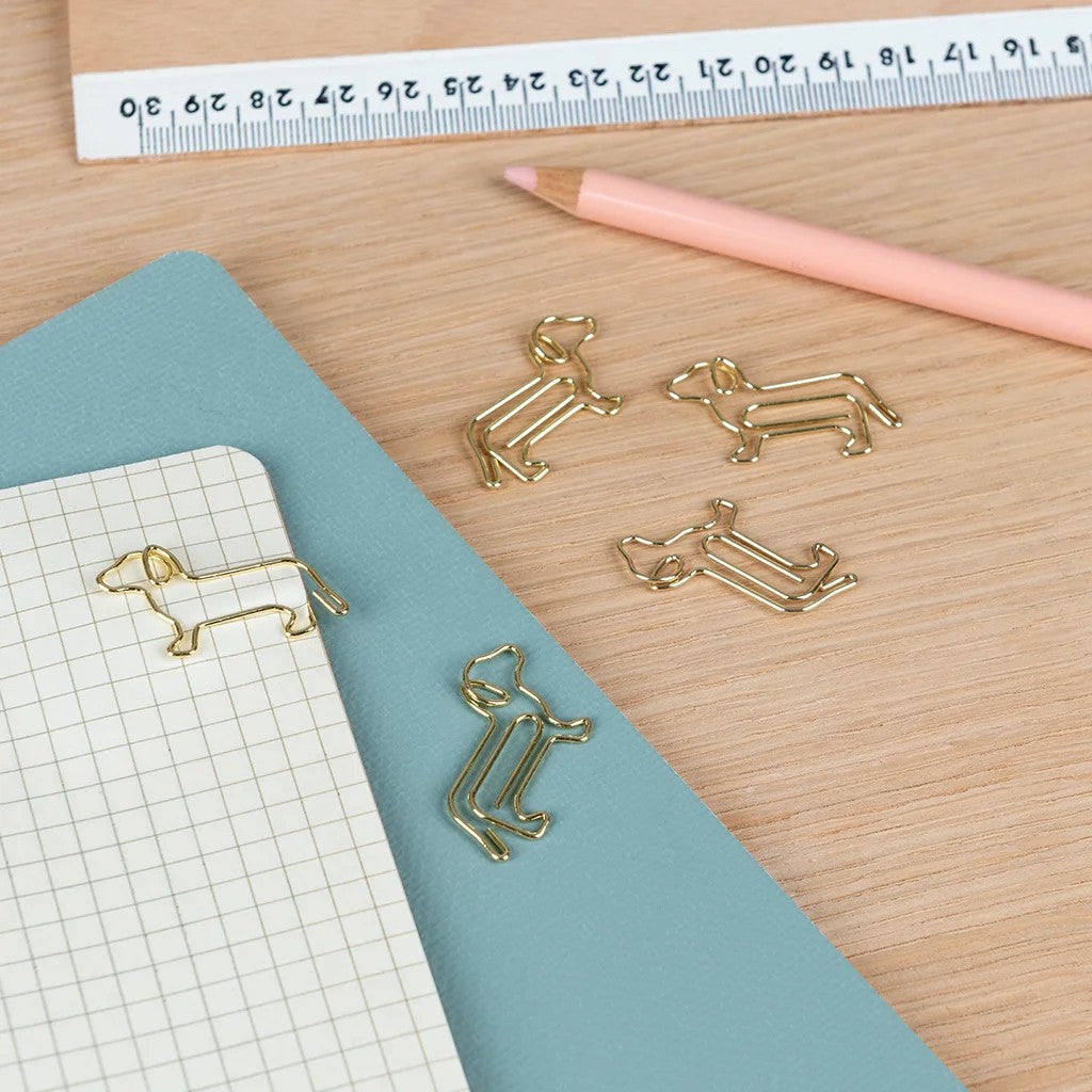Goudkleurige hondvormige paperclips op een notitieblok met ruitjes naast een blauwgroen omslag, een roze potlood en een liniaal op een houten bureau