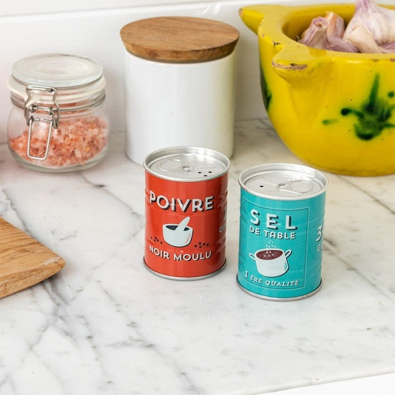 Rode peperstrooier en turquoise zoutstrooier in blikvorm op een marmeren aanrecht, naast een glazen pot met roze zout en een gele vijzel met knoflook