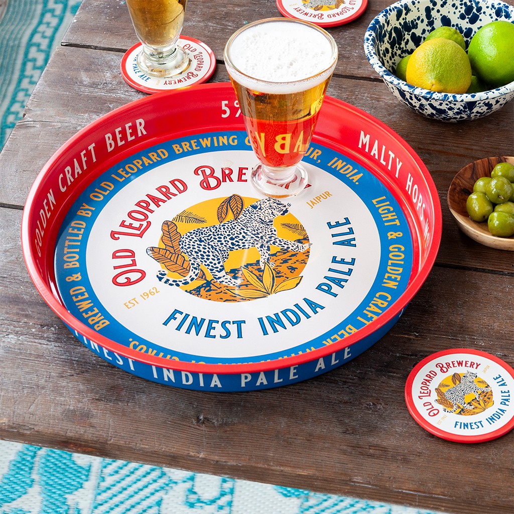 Rond dienblad met rode rand en luipaardprint op een houten tafel, met een glas bier op het dienblad, een kom limoenen en een houten schaaltje olijven ernaast