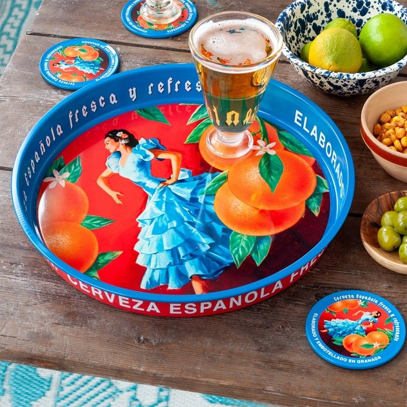 Blauw dienblad met flamencodanseres en sinaasappels op een houten tafel, met een glas bier, schaaltjes met limoenen en noten en onderzetters ernaast
