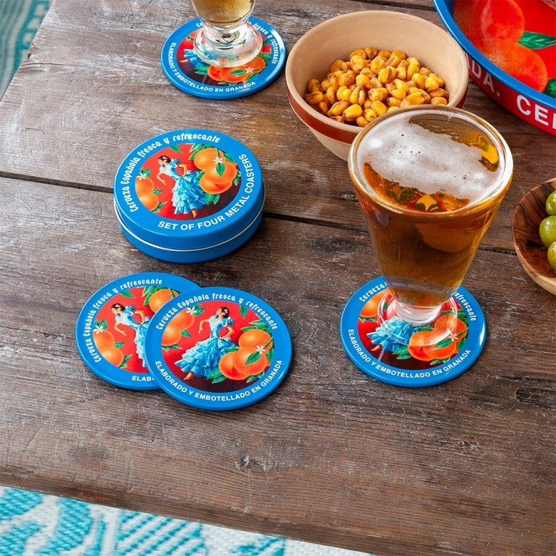 Blauwe onderzetters met danseres op een houten tafel, met een glas bier op een onderzetter en een schaal gekruide noten en olijven ernaast