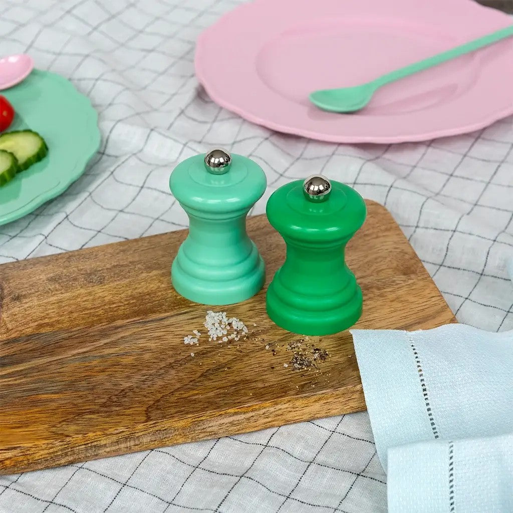 Twee groene molens in een donkere en een lichte tint met zilverkleurige bol op een houten plank, met uitgestrooid zout en peper, op een geruit kleed met een roze bord en een turquoise lepel
